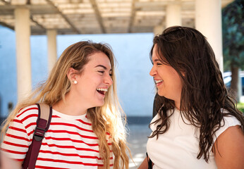 Two college girls smiling and laughing together in a sunny university campus environment with...