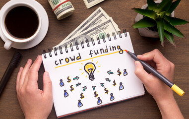Business concept. Woman hand writing in notebook crowdfunding sign on table with dollars, panorama