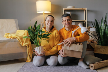 A cheerful couple in yellow tops sits on the floor of their new home, holding a houseplant and a...
