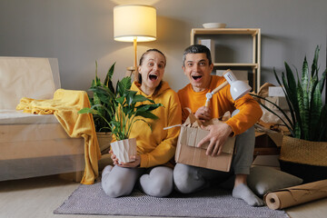 A delighted couple sits on the floor of their new home, holding a houseplant and a box of items,...