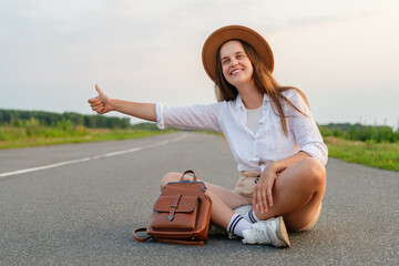 Young woman in a hat and white shirt sitting on a rural road, smiling and hitchhiking with her thumb up.
