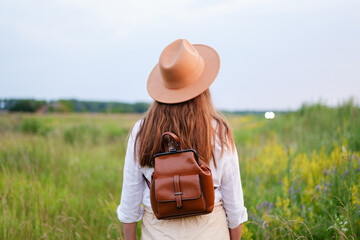Woman with a backpack and hat standing in a field, enjoying nature and summer freedom from behind...