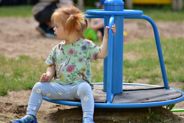 Toddler girl sits on a blue merry-go-round at a park, holding the handle and looking sideways,...