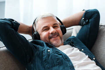 Fototapeta premium Happy matured man reclining on couch at home, listening to music and smiling, using wireless modern headset, closeup, copy space. Modern technologies and entertainment concept
