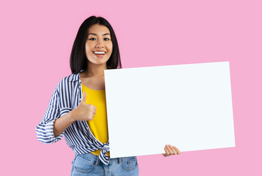 Place For Your Ad. Smiling asian woman holding empty blank board and showing thumbs up sign gesture isolated on pink studio background. Happy lady standing with white square paper for template - Powered by Adobe