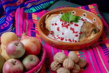 Classic Mexican chile en nogada served in a clay dish with fresh ingredients: apple, pear, walnut, and pomegranate over a vibrant textile background.
