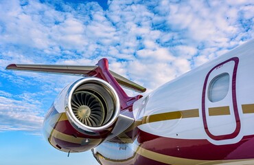 A polished private jet engine with visible turbine blades and rear fuselage, including the emergency exit door and T-tail assembly. The aircraft is parked under a partly cloudy sky. 
