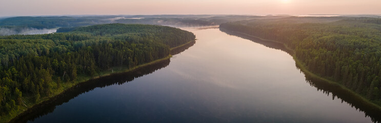 Aerial drone panorama of a calm northern lake at sunrise with mist on the water and surrounding forest
