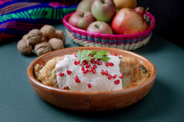 Classic Mexican chile en nogada served in a clay dish with fresh ingredients: apple, pear, walnut, and pomegranate over a vibrant textile background.
