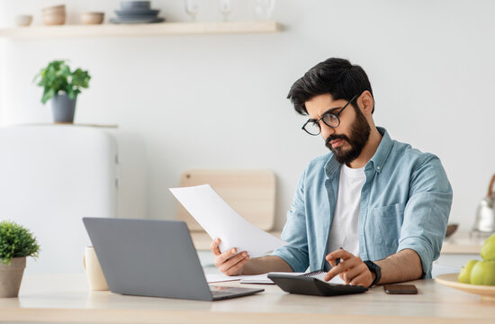 Paying bills, taxes at home online. Young arab man using calculator and laptop computer, calculating taxes or planning budget while sitting at kitchen table at home, copy space