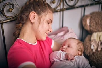 Young blonde woman cradles sleeping baby girl with light hair. Warm lighting, soft background...