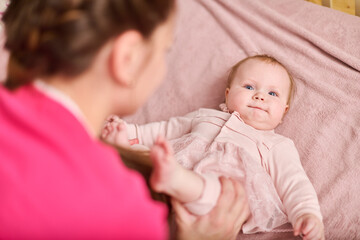 Smiling adult woman with brown hair interacts with baby girl. Baby lies on pink blanket. Warm...