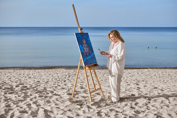 Young woman artist stands near ocean, painting on easel. Serene beach environment complements her...