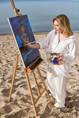 Young woman artist painting mystical portrait on canvas at serene beach. Artwork depicts surreal...