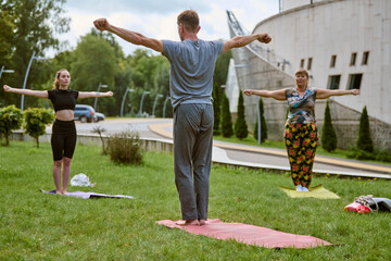 Three adults practicing yoga on mats in park. Sunny day with green grass, trees, and modern...