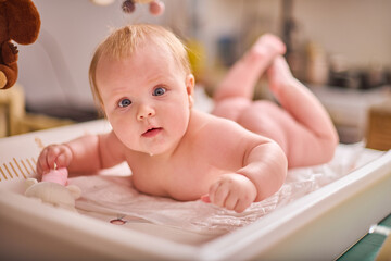 Blonde baby girl on changing table, playful mood. Soft lighting, warm colors, cozy room. Cute toys,...