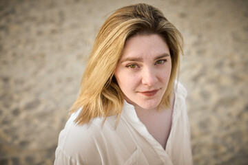 Young woman with blond hair gazes softly towards camera, standing on sandy beach. Her white shirt...