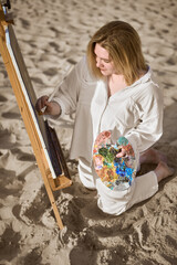 Young woman artist in white shirt creates masterpiece on easel at sandy beach. Soft morning light...