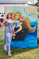 Young female artist with brown hair stands proudly by colorful mermaid painting. Daylight enhances...