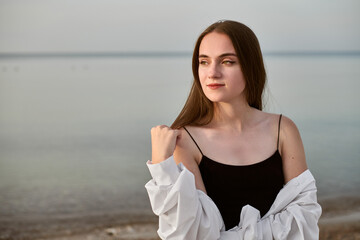 Young woman in black dress and white shirt stands contemplatively by serene sea. Her relaxed pose...