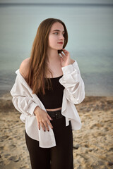 Young woman stands on sandy beach, wearing white shirt over black outfit, gazing contemplatively at...
