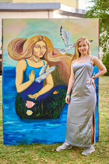 Young female artist, blonde, caucasian, poses proudly with her vibrant painting in grassy outdoor...
