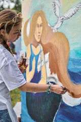 Young woman with long hair painting mural outdoors. Focused expression, holding paintbrush....