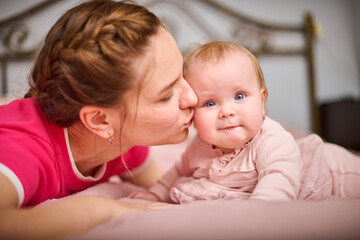 Young mother with braided hair kisses light-haired baby girl on pink bedspread, expressing warmth...