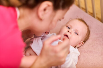 Young mother with brown hair engages with joyful baby girl. Baby lies in crib, smiling brightly....