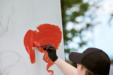 Young female artist in black cap painting with red brush strokes on large canvas. Trees and blurred...