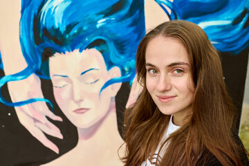 Young adult caucasian female with long brown hair smiling beside colorful mural. Artwork behind her...