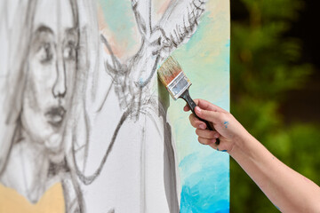 Close-up of female hand painting canvas outdoors, sketching woman face. Natural lighting highlights...