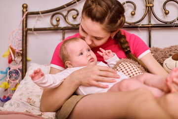Young woman with braided brown hair cradles fair-skinned baby girl on bed. Warm lighting creates...