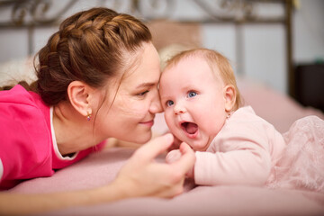 Young woman with brown hair and smiling baby girl on pink bed. Light-hearted interaction, creating joyful atmosphere. Warm lighting enhances bond