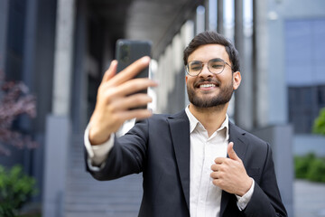 A smiling businessman in a suit takes a selfie with a smartphone and gives a thumbs-up outside a...