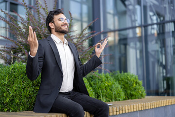 A smiling businessman in a suit meditates outdoors, showcasing a moment of tranquility and balance in a modern urban environment.