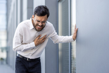 A man in business attire clutches his chest, likely experiencing chest pain, reflecting possible...