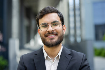 A smiling businessman with a beard and glasses stands confidently outdoors in front of a building,...