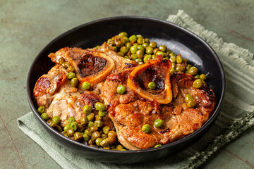 Beef, veal ossobuco with green peas, onion in a black plate and dark green table with runner. Italian meat and vegetable meal.