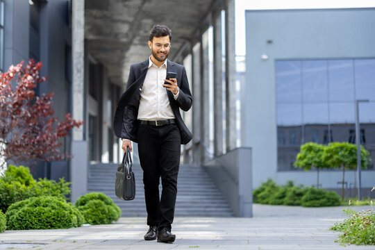 A smiling businessman in a suit walks while looking at his phone near a modern building with greenery and holding a briefcase. - Powered by Adobe