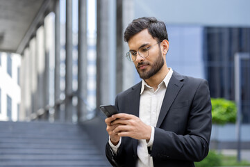 An Indian businessman in a suit checks his phone outside a modern building. He is wearing glasses,...