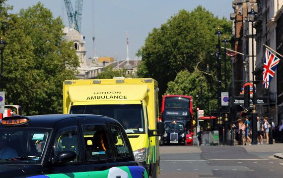 Urban perspective along a busy street in central London with taxi, yellow ambulance. Defocus background. with red bus and English flag at a building - Powered by Adobe