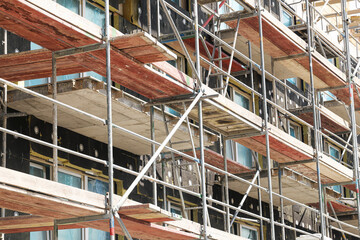 Building under construction scaffolding. Metal frame structure supports multi floor facade. Apartment complex renovation with red planks. Industrial site with safety rails and modern windows exposed