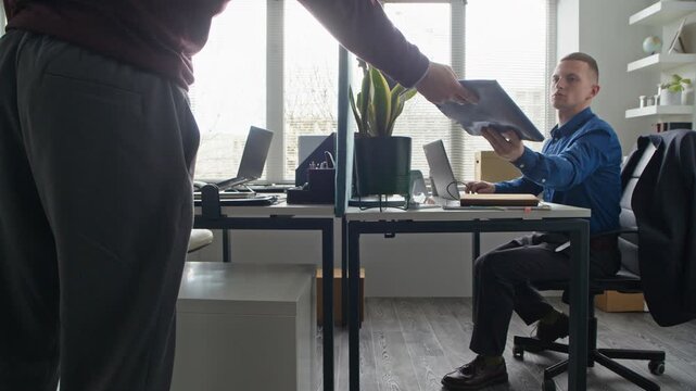 Side view of adult man handing folder with documents to male colleague sitting nearby at desk then returning to work on laptop in office workspace