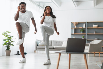 Obraz premium Online Family Workout. African American Husband And Wife Training Together In Living Room, Doing Twist Knees To Elbow Exercise. Happy Black Standing And Lifting Leg Up, Using Laptop, Watching Tutorial