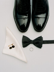 Formal attire laid out with polished shoes, bow tie, cufflinks, and wedding ring on white background
