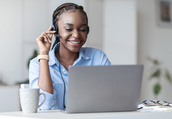 Customer Support Concept. Black woman manager wearing headset and using laptop in office, consulting clients, sitting at desk at workplace, free space