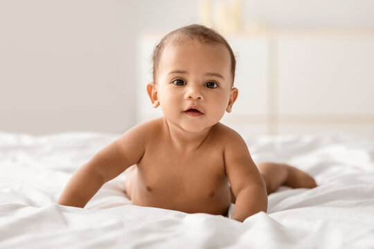 Closeup portrait of sweet naked black baby crawling on bed on the white bedsheets, indoors. Cute positive little afro infant lying on belly, looking at camera. Free space, blurred background - Powered by Adobe