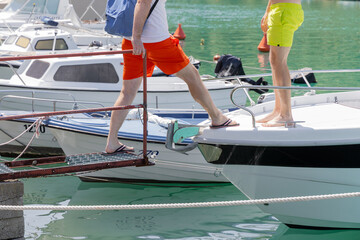 Two individuals in bright swimwear stepping onto a boat from a dock, enjoying a sunny day at the marina with vibrant water reflections
