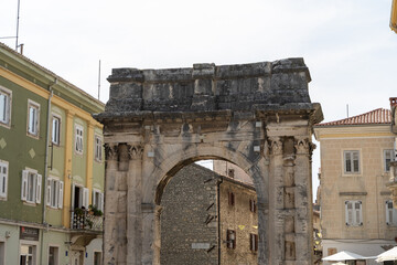 Obraz premium Pula, Croatia - July 15, 2025: Ancient stone archway stands proudly amidst colorful buildings in a historic town square with vibrant atmosphere